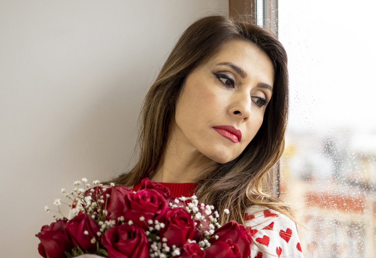 kobieta w wieku 35 plus z bukietem róż stoi przy oknie zamyślona i smutna