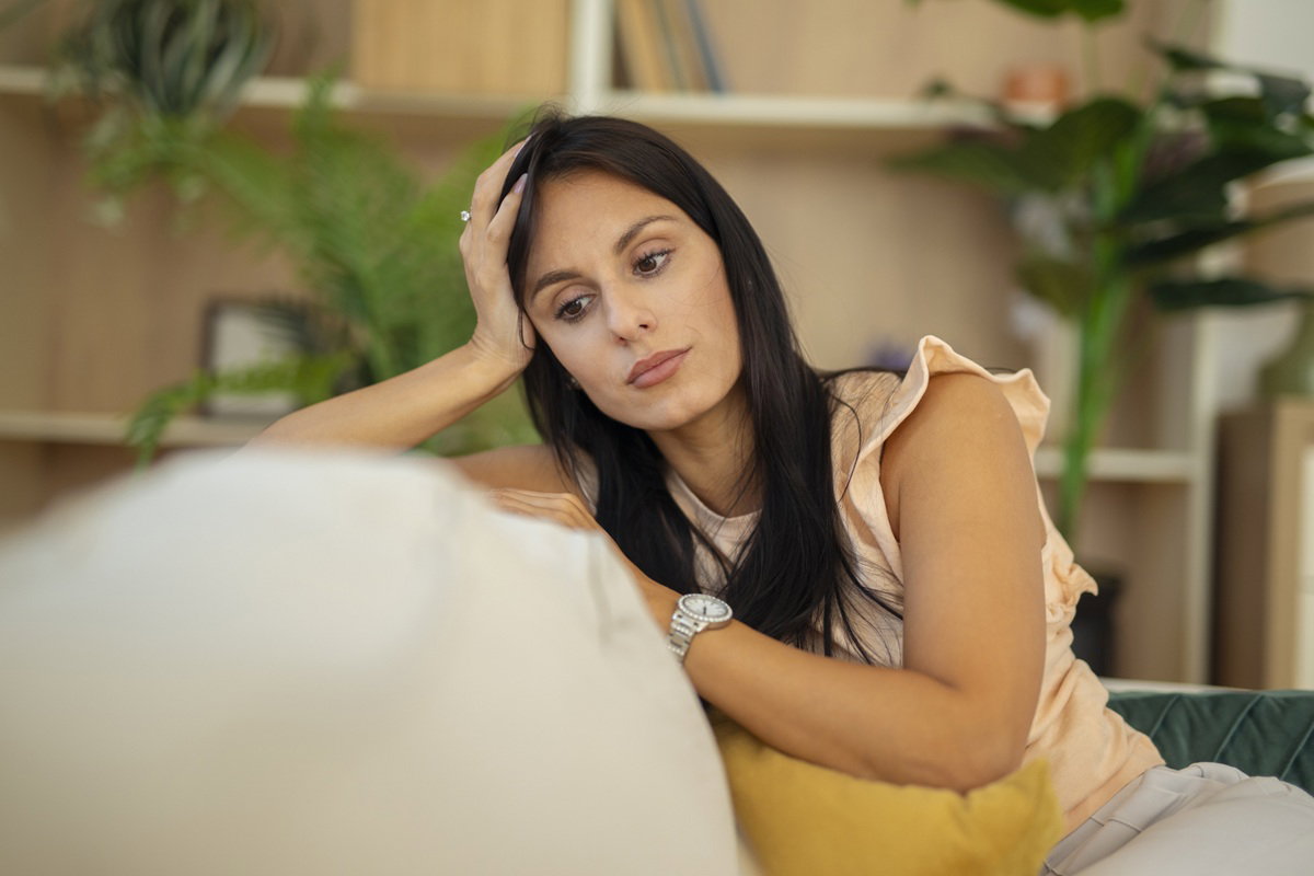 kobieta w wieku 30 plus z czarnymi długimi włosami siedzi na kanapie zamyślona i smutna