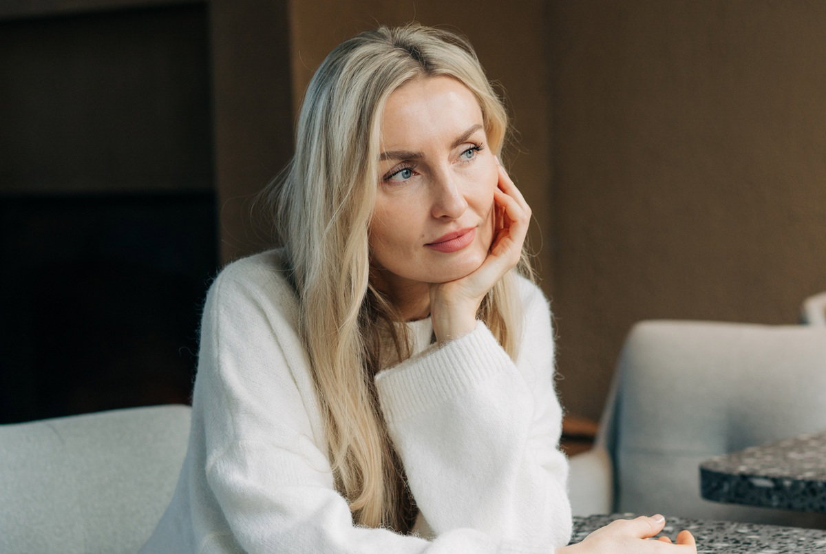kobieta blondynka w wieku 35 plus w jasnym swetrze siedzi zamyślona i smutna w kawiarni