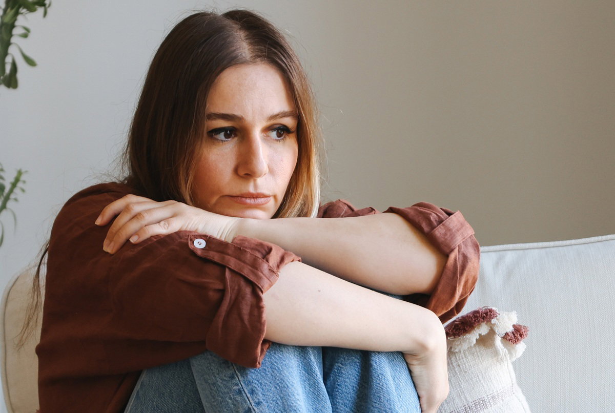 kobieta w wieku 30 plus w brązowej koszuli siedzi na kanapie zamyślona i smutna