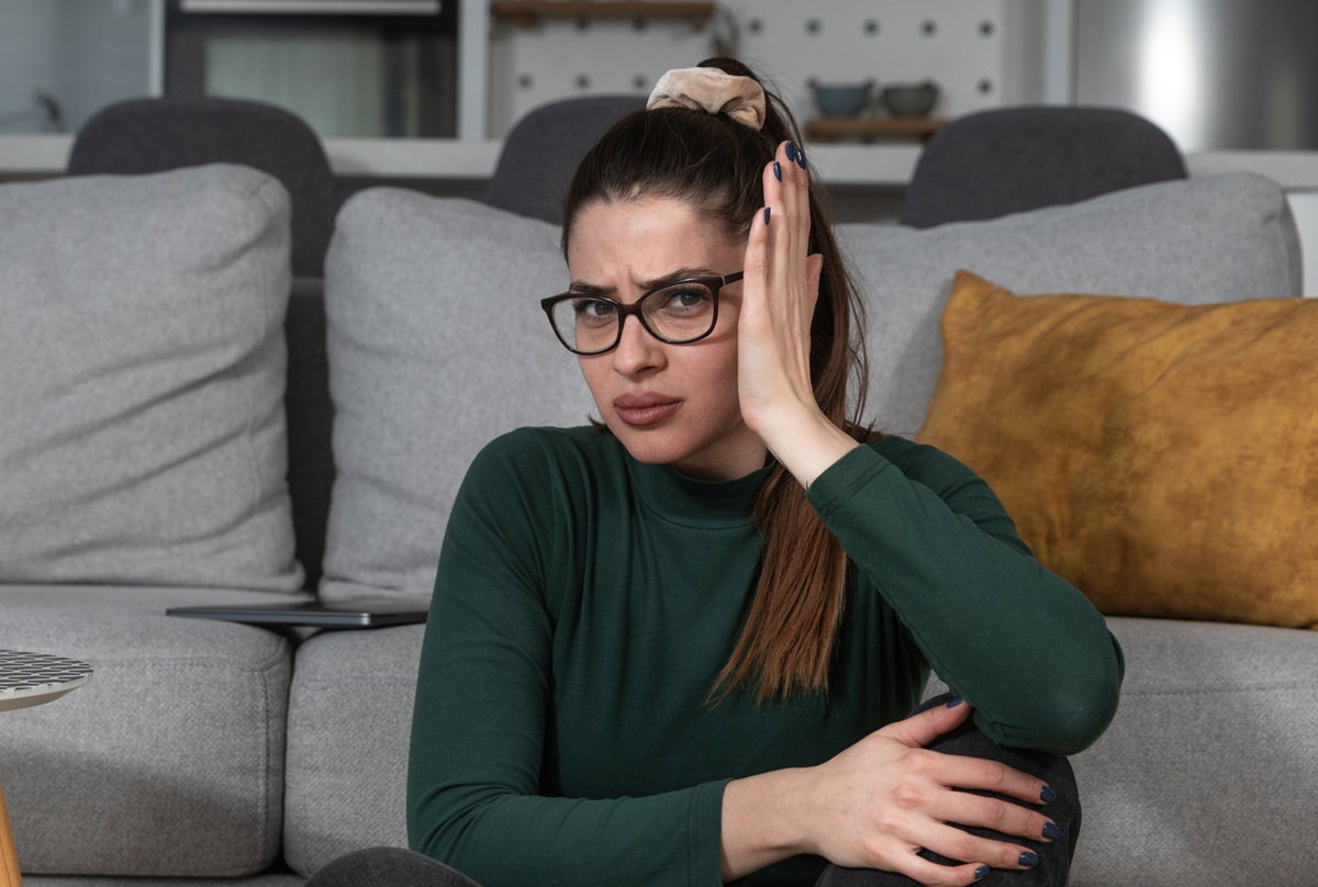 kobieta w wieku 25 plus w zielonej bluzce i okularach siedzi przy kanapie  wyraźnie zdegustowana