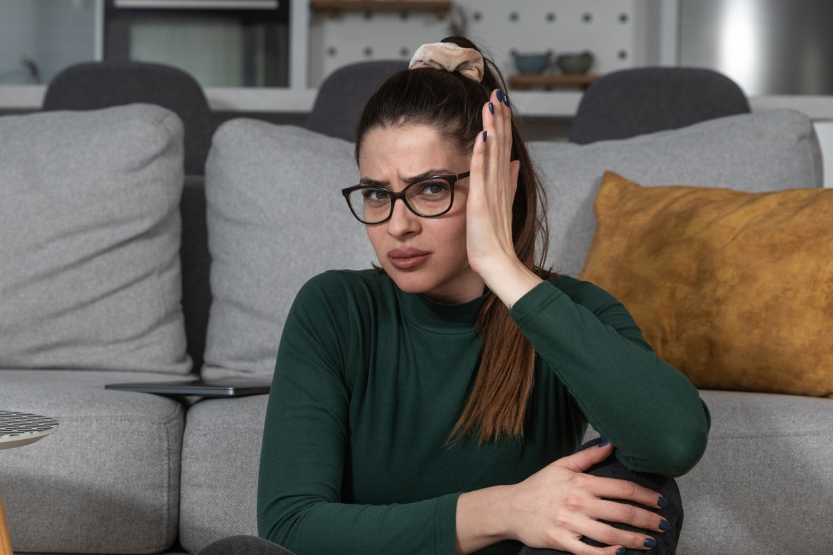kobieta w wieku 25 plus w zielonej bluzce i okularach siedzi przy kanapie wyraźnie zdegustowana