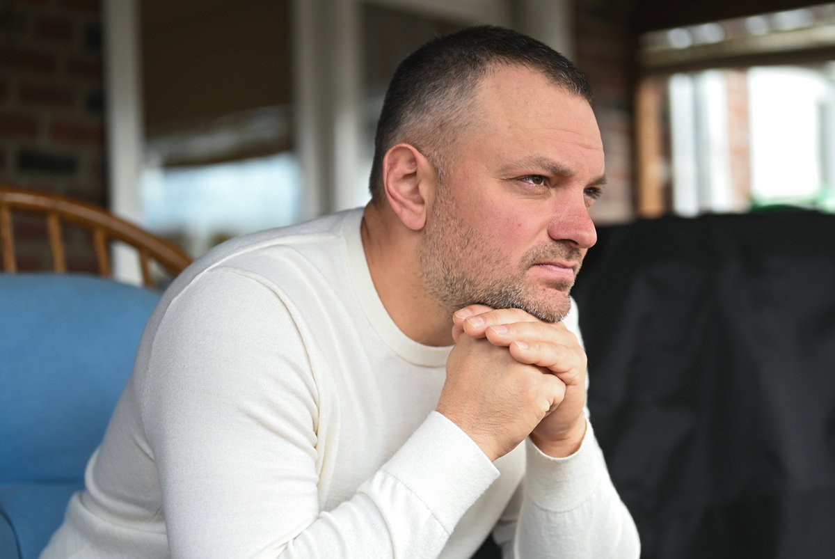 mężczyzna w wieku 35 plus z zarostem i w białym longsleevie siedzi zamyślony w fotelu