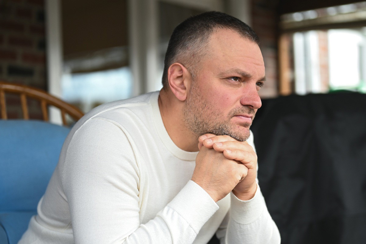 mężczyzna w wieku 35 plus z zarostem i w białym longsleevie siedzi zamyślony w fotelu