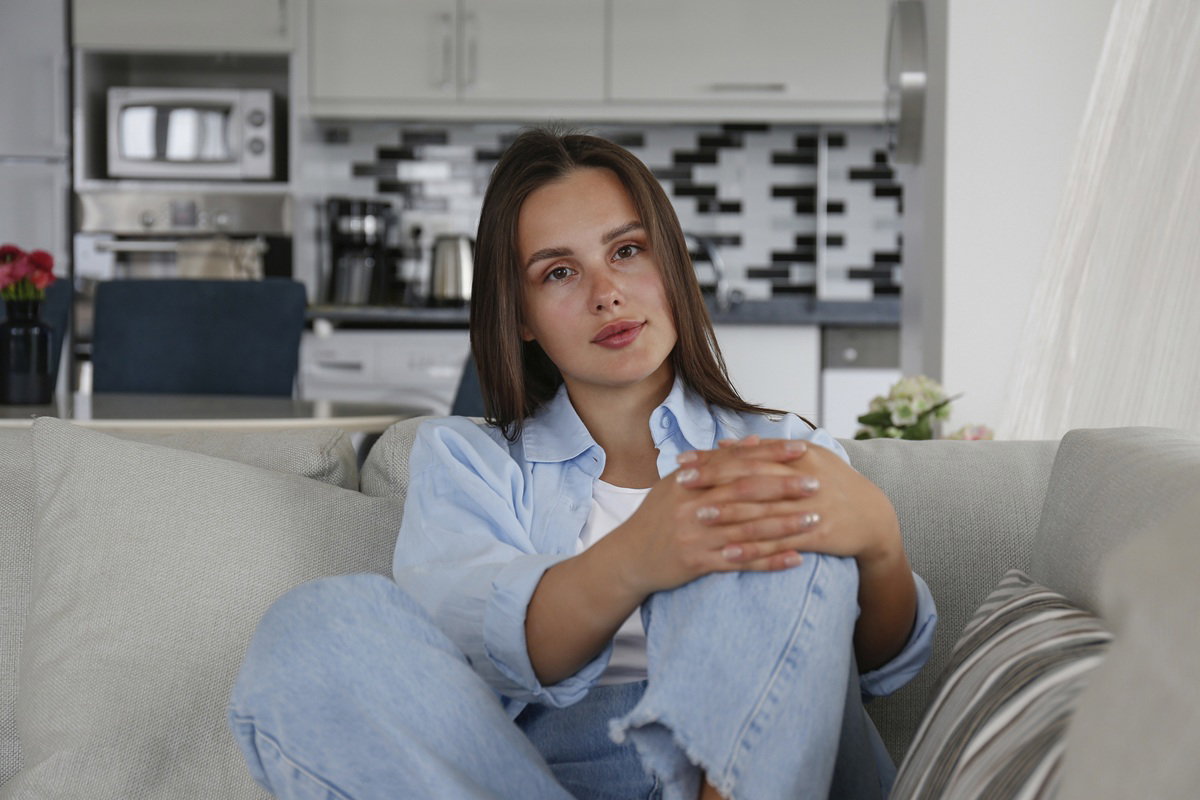 kobieta w wieku 30 plus  w jasnej koszuli siedzi na kanapie na tle kuchni zamyślona