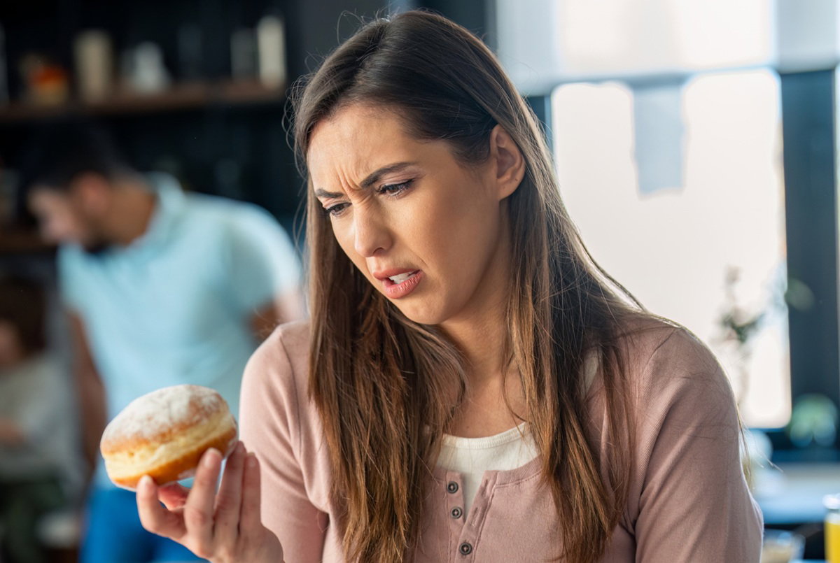 kobieta w wieku 30 plus z pączkiem w ręku stoi w kuchni i krzywi się z niesmakiem
