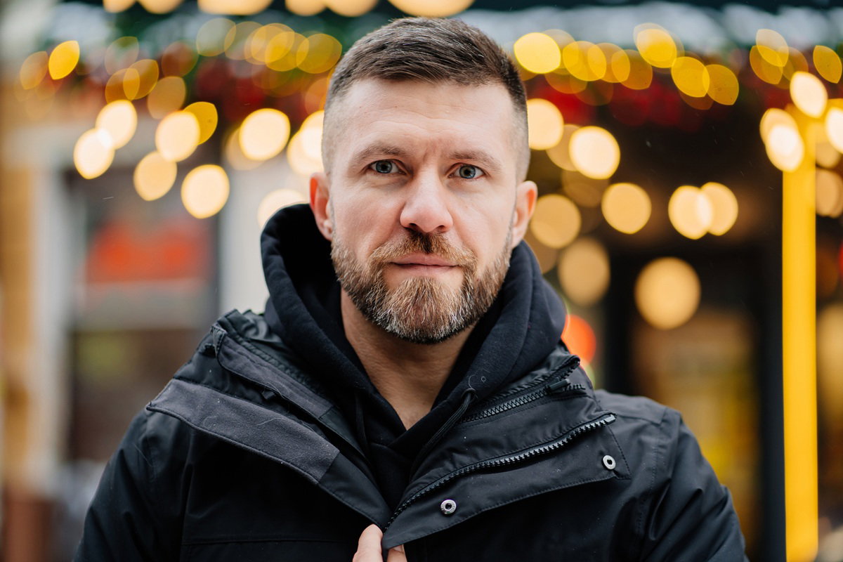 mężczyzna w wieku 30 plus z wąsami i brodą stoi na tle ciepłych lampek z lekko smutną miną