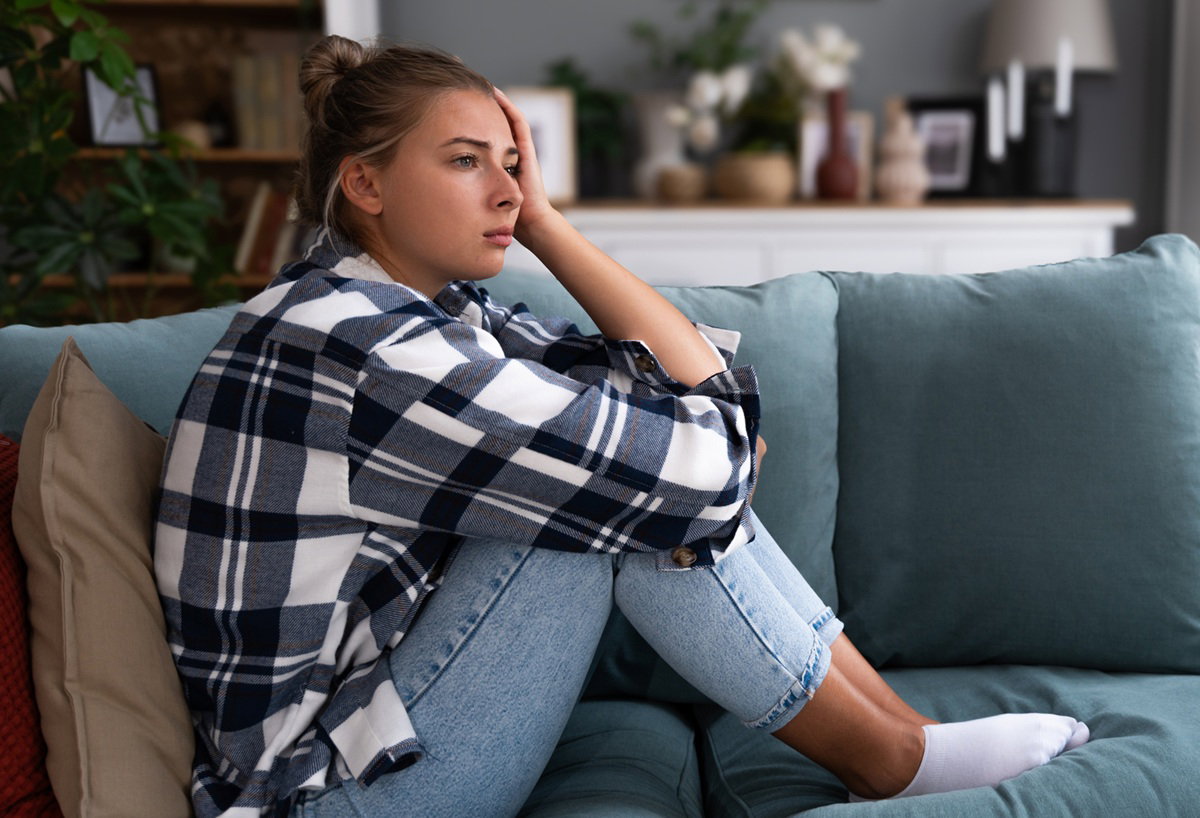 kobieta w wieku 25 plus w kraciastej koszuli siedzi na kanapie zamyślona na tle kuchni