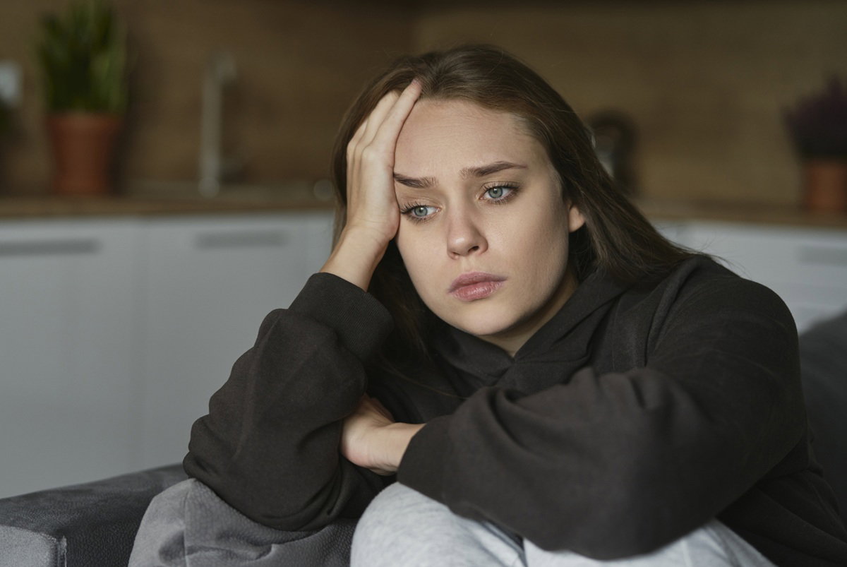 kobieta w wieku 25 plus w ciemnej bluzie siedzi na kanapie na tle kuchni smutna i zamyślona