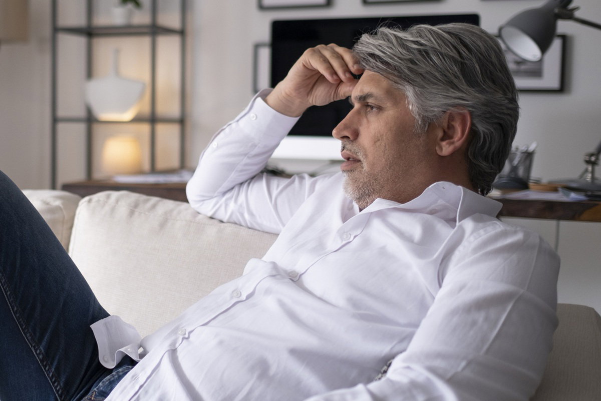 mężczyzna w wieku 40 plus w białej koszuli siedzi zamyślony na kanapie