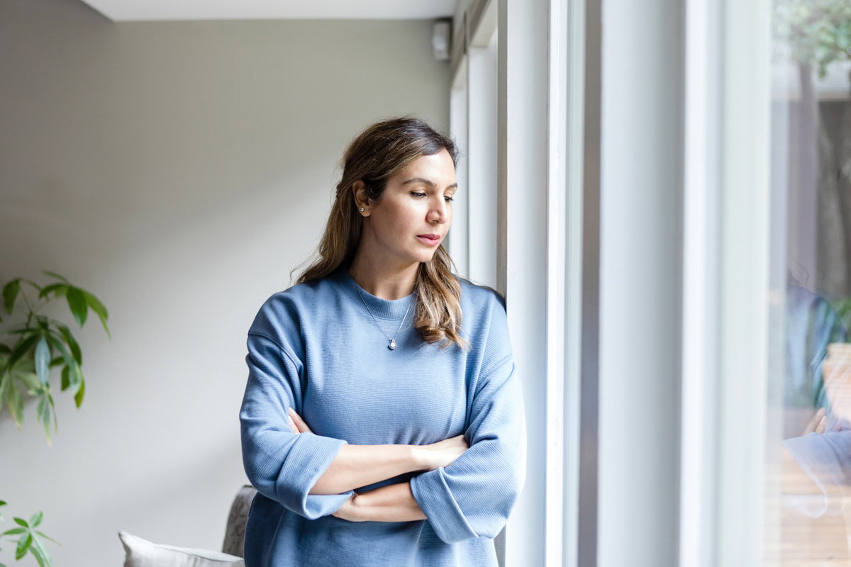 kobieta w wieku 40 plus w niebieskiej bluzce stoi przy oknie smutna i zamyślona