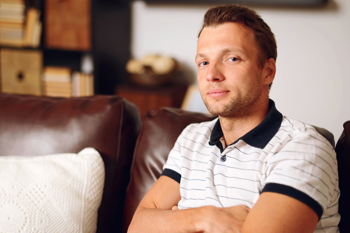 mężczyzna w wieku 30 plus w koszulce polo siedzi na kanapie z lekko kpiącym uśmiechem