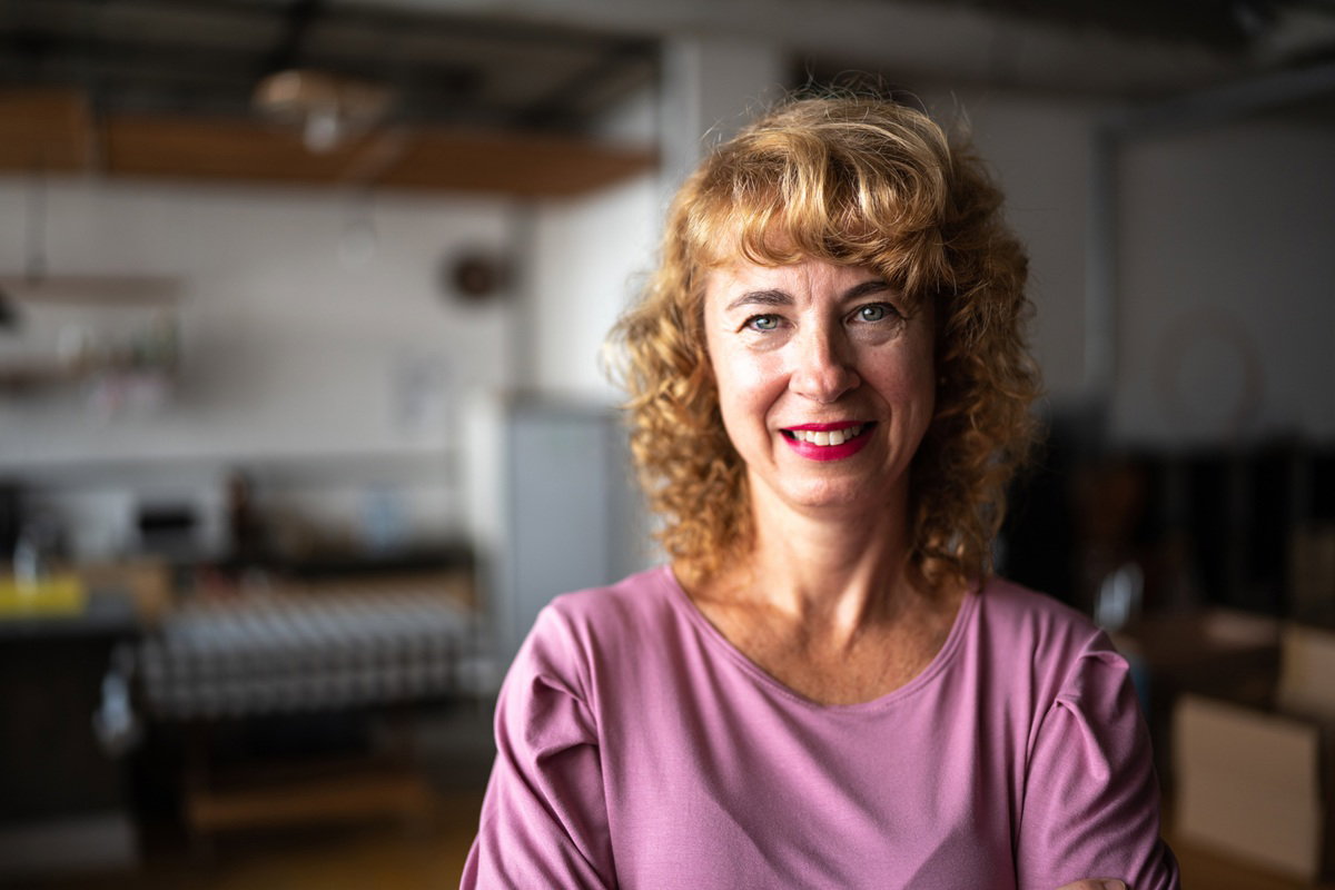 kobieta w wieku 50 plus w różowej bluzce stoi w mieszkaniu i uśmiecha się rozpromieniona