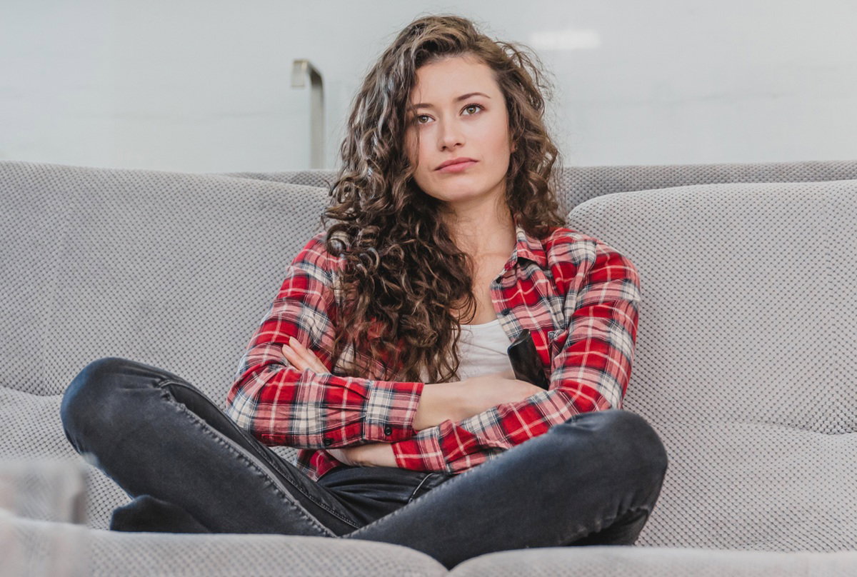 kobieta w wieku 30 plus kręcone długie włosy i w kraciastej koszuli siedzi na kanapie wkurzona