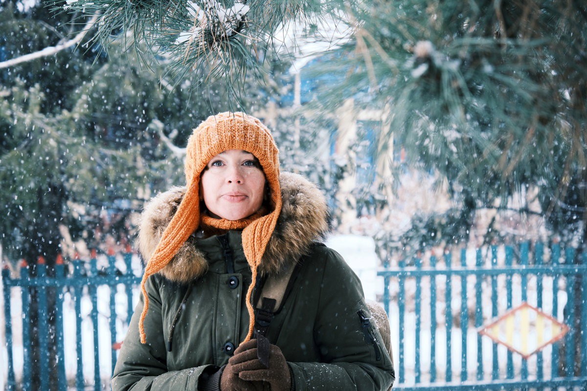 kobieta 30 plus w pomarańczowej czapce i zielonej kurtce stoi na zewnątrz na tle zaśnieżonego pejzażu