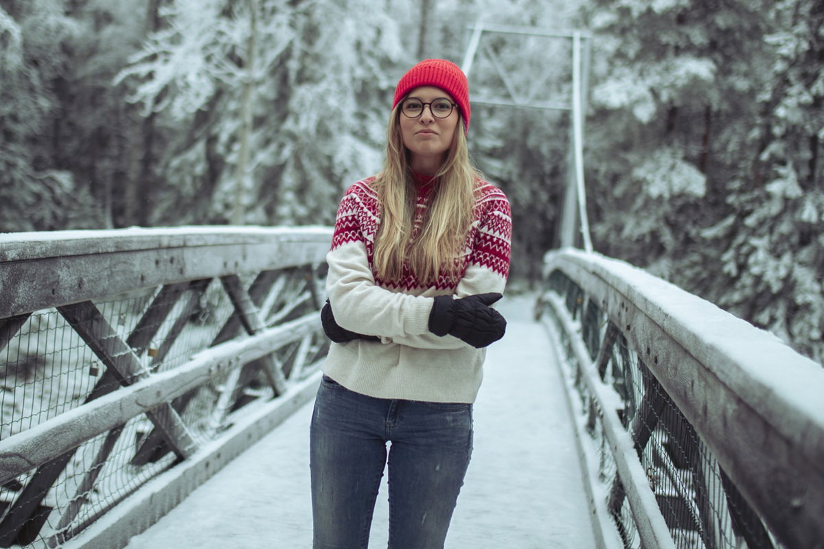 kobieta w wieku 30 plus w okularach w swetrze i czerwonej czapce i rękawiczkach stoi na tle zmowego pejżażu dumna