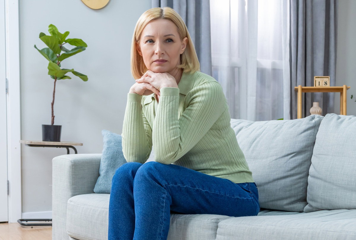 kobieta w wieku 40 plus w zielonym sweterku siedzi na kanapie zamyślona i smutna