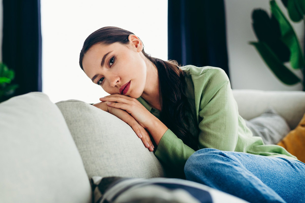kobieta w wieku 30 plus z długimi ciemnymi włosami siedzi na kanapie zamyślona i smutna