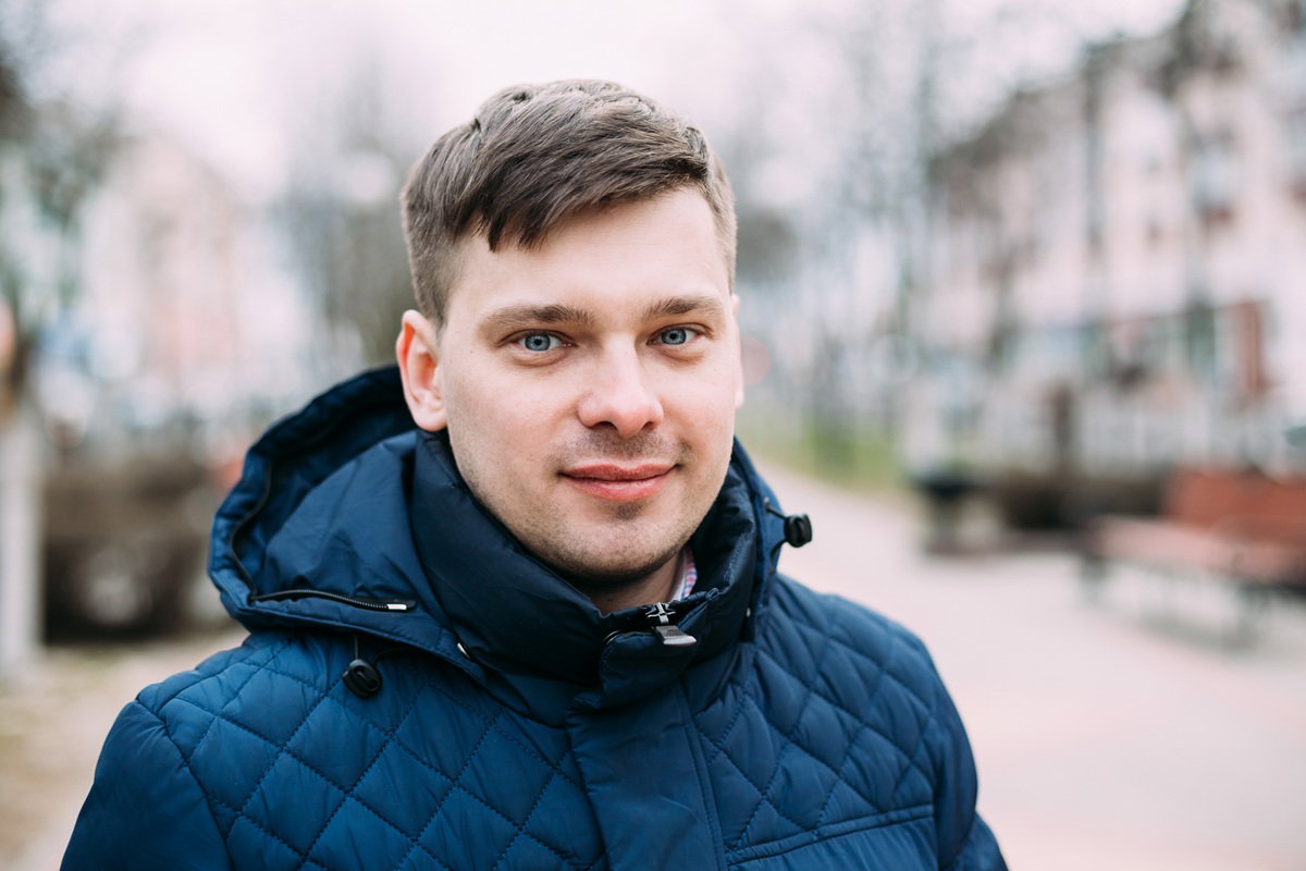 mężczyzna w wieku 30 plus w granatowej kurtce stoi na tle osiedla i lekko się uśmiecha