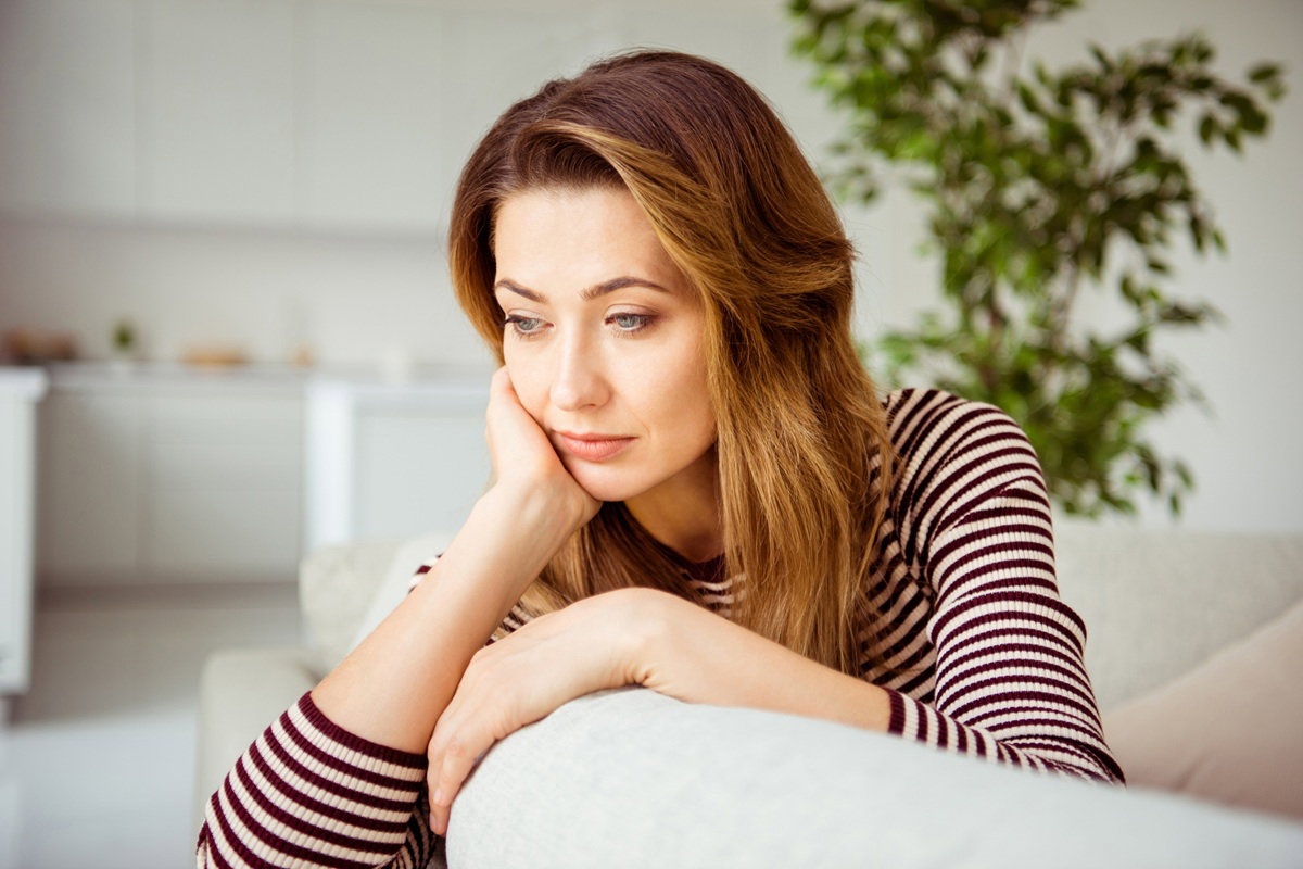 kobieta w wieku 35 plus w bluzce w paski siedzi na kanapie na tle kuchni zamyślona