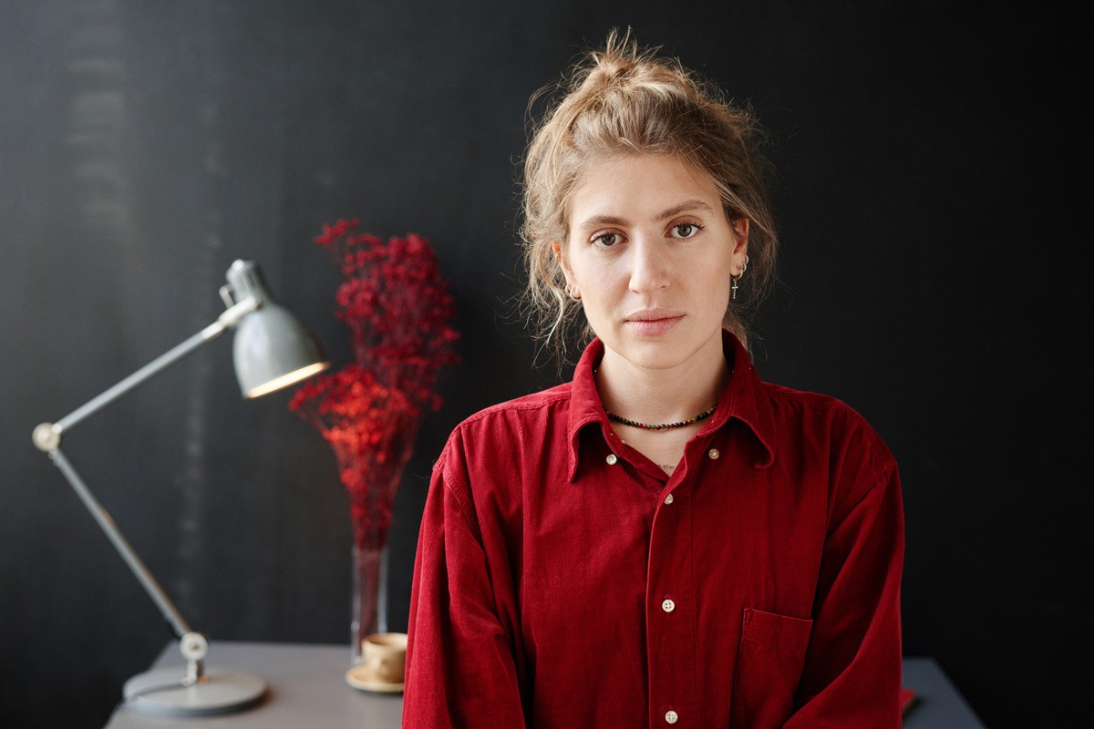 kobieta w wieku 25 plus w czerwonej koszuli stoi zamyślona na tle szarego wnętrza