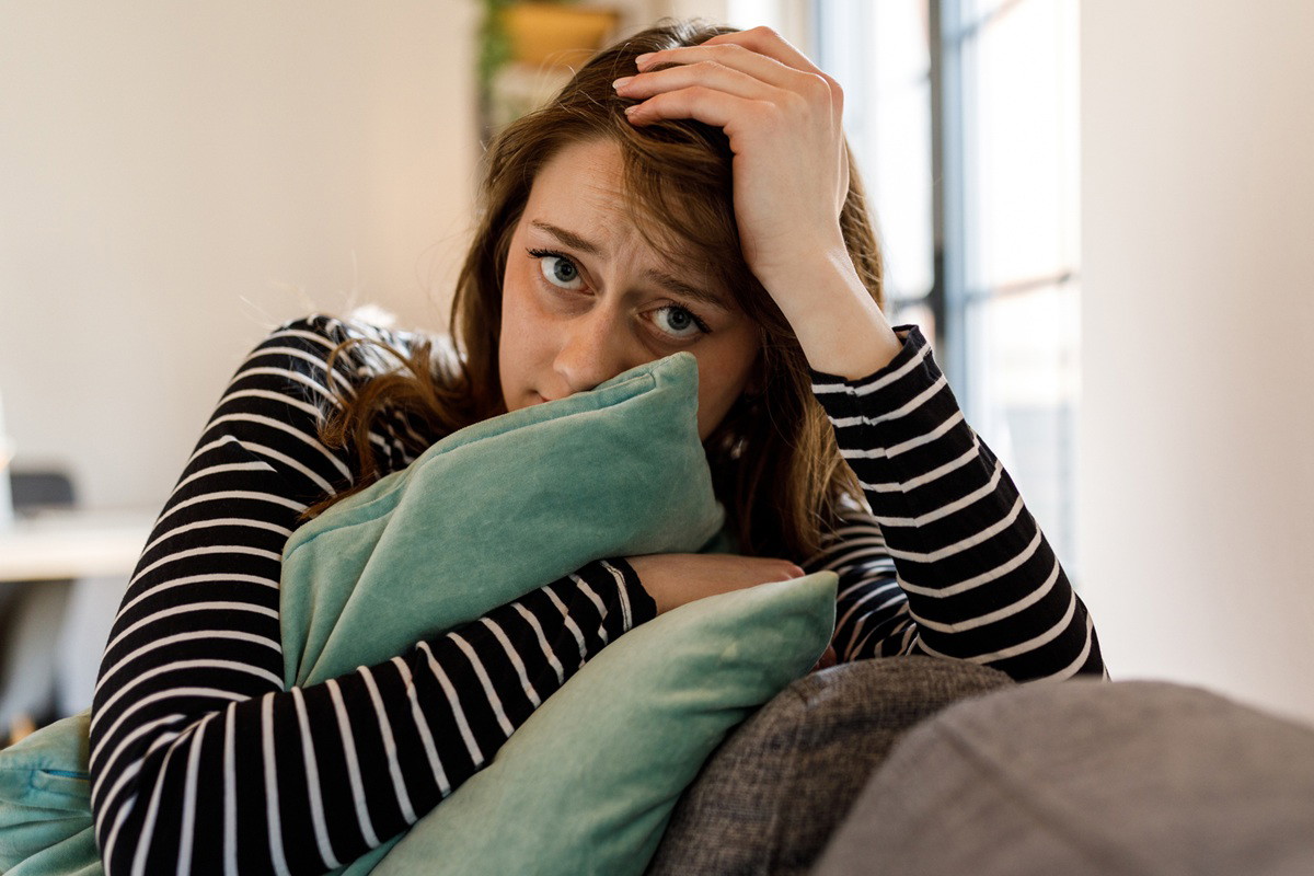 kobieta w wieku 25 plus w bluzce w paski siedzi na kanapie i przyciska poduszkę przerażona