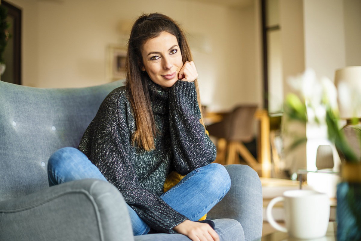 kobieta w wieku 30 plus z długimi włosami i w melanżowym swetrze siedzi na sofie i lekko się uśmiecha