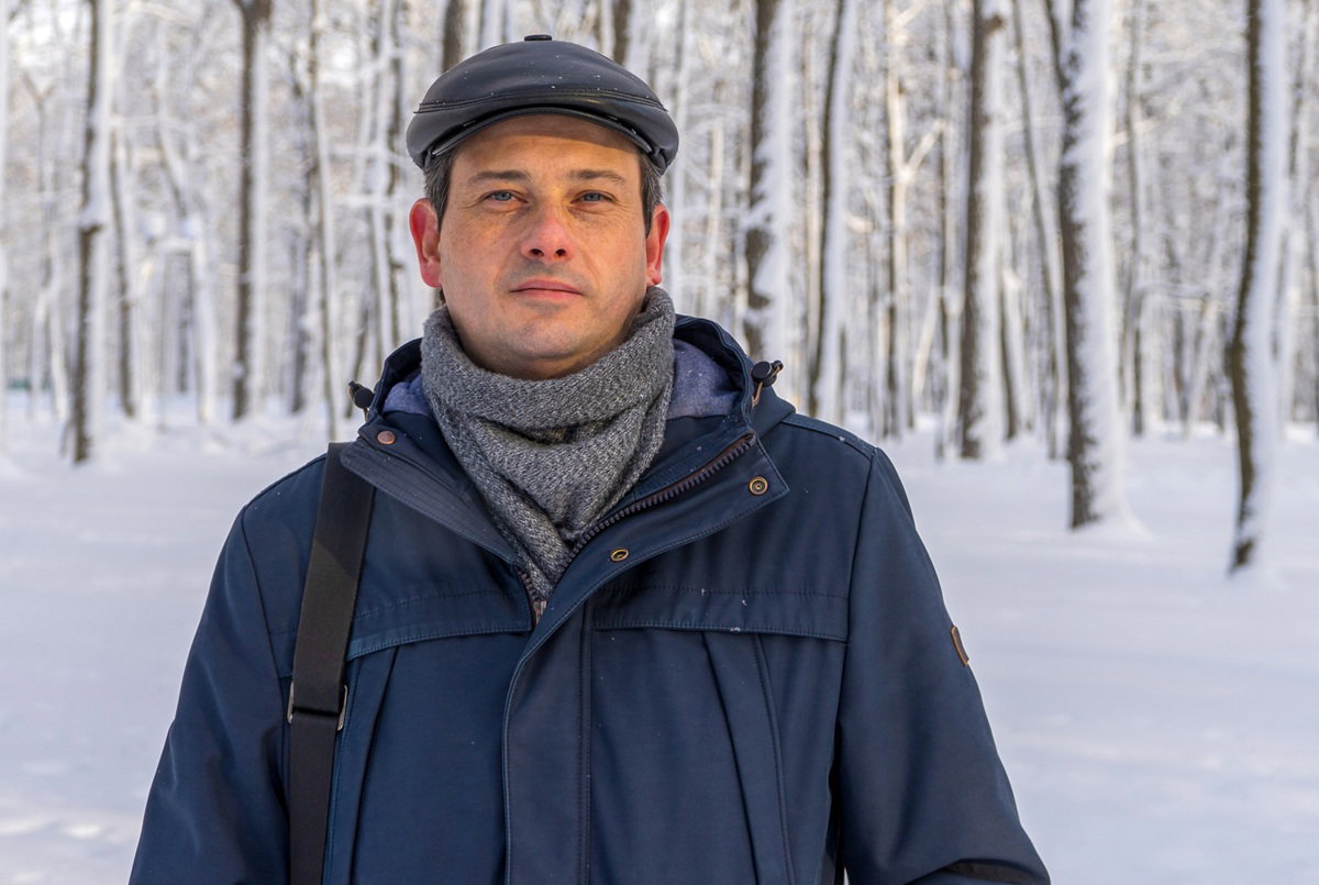 mężczyzna w wieku 35 plus w skórzanym kaszkiecie stoi z torbą i w granatowej kurtce na tle zimowego lasu