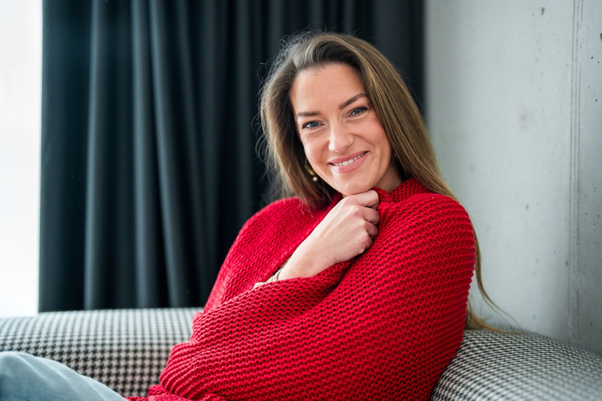 kobieta w wieku 40 plus siedzi owinięta w czerwonym kocu na kanapie i zalotnie się uśmiecha