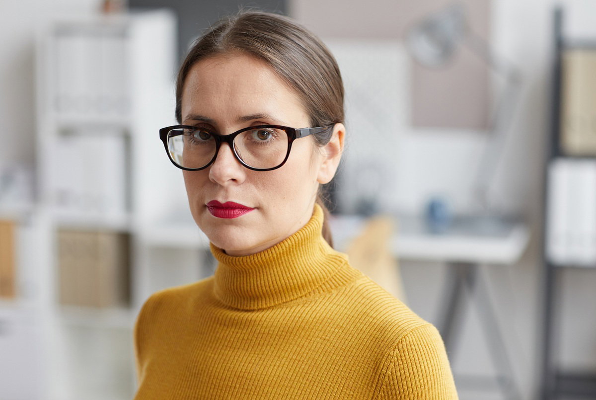 kobieta w wieku 35 plus w okularach w żółtym golfie stoi w biurze z poważną miną