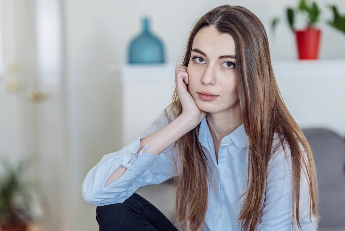 kobieta w wieku 20 lat z długimi włosami siedzi na kanapie w salonie w jasnej koszuli zamyślona