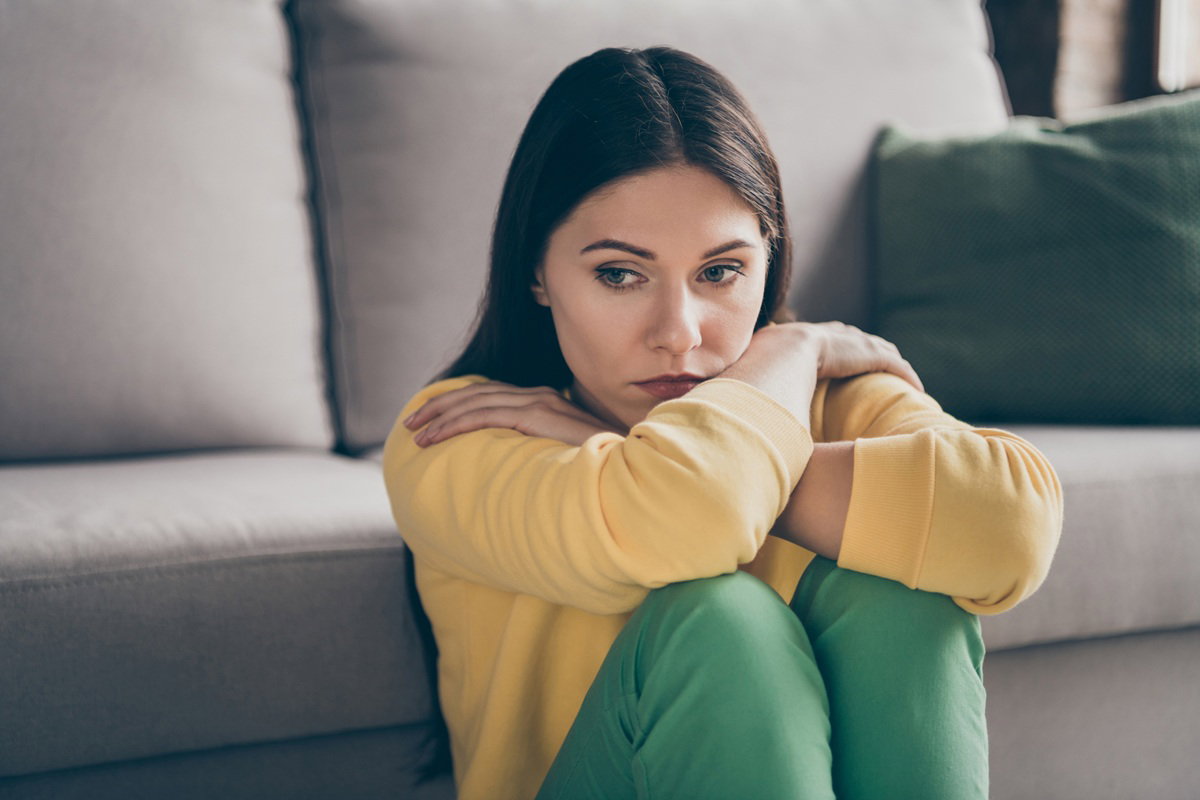 kobieta w wieku 30 plus w żółtej bluzie siedzi na podłodze przy kanapie zamyślona