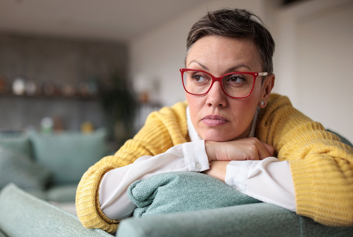kobieta w wieku 45 plus w okularach i żółtym swetrze leży na kanapie zamyślona