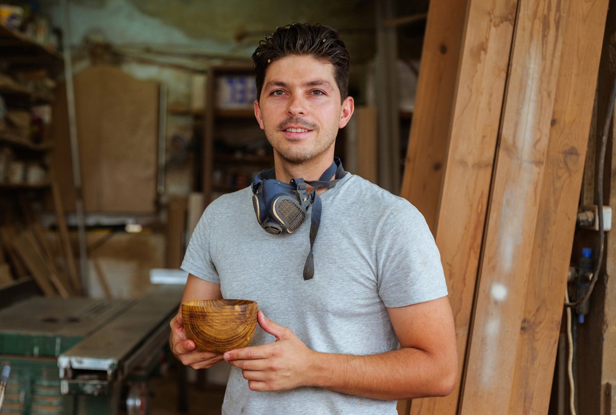 mężczyzna w wieku 30 plus stolarz z drewnianym naczyniem w dłoniach stoi na tle stolarni