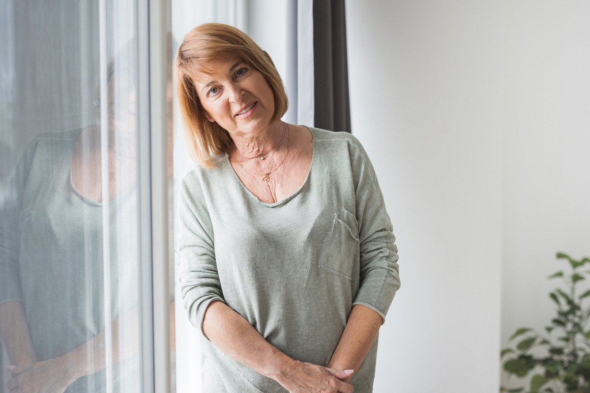 kobieta w wieku około 60 lat w jasnej bluzce stoi przy oknie zamyślona