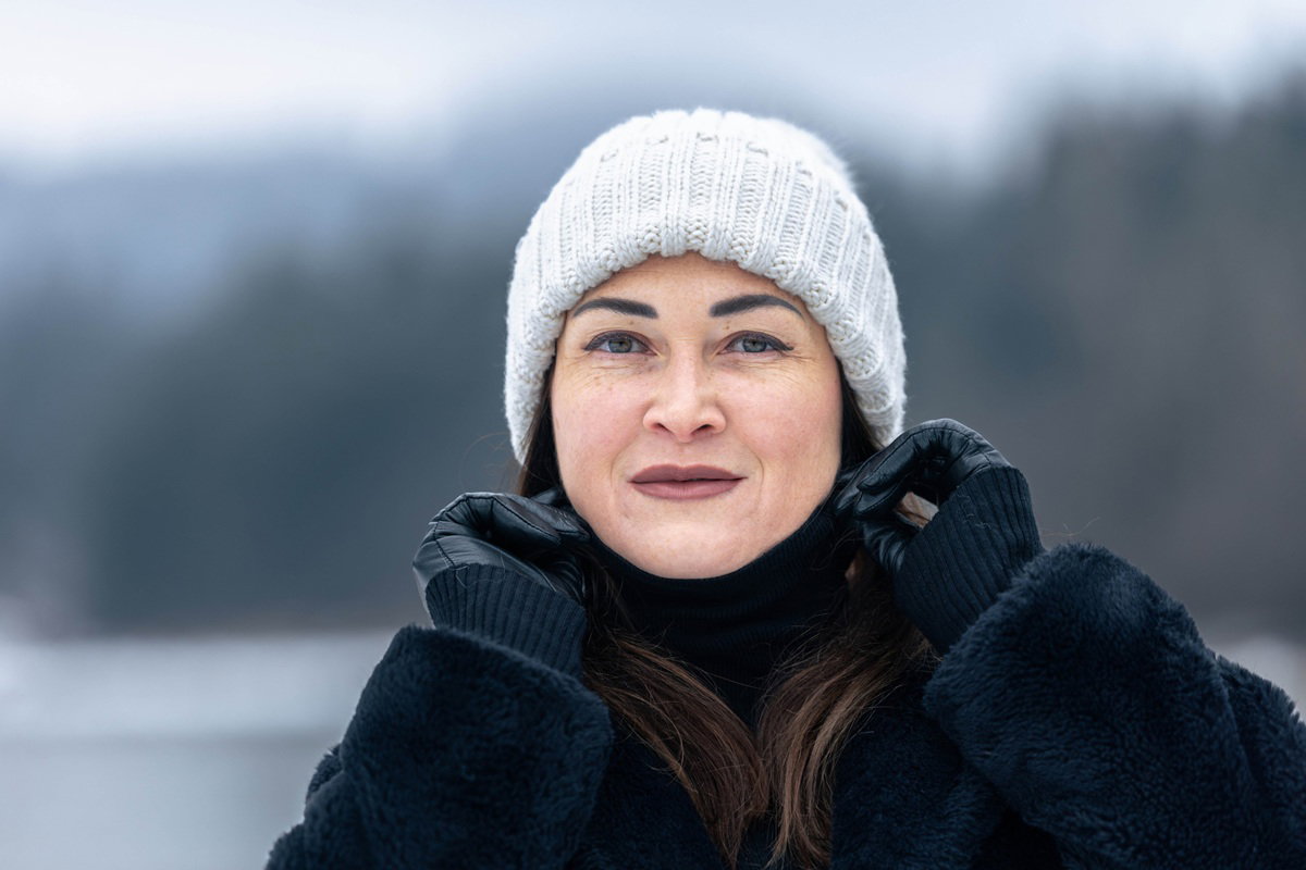 kobieta w wieku 45 plus w czapce i czarnym futerku stoi na zewnątrz i lekko się uśmiecha
