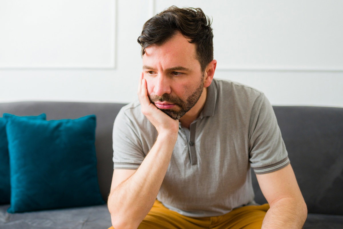 mężczyzna w wieku 35 plus z ciemnym zarostem siedzi na kanapie zmartwiony