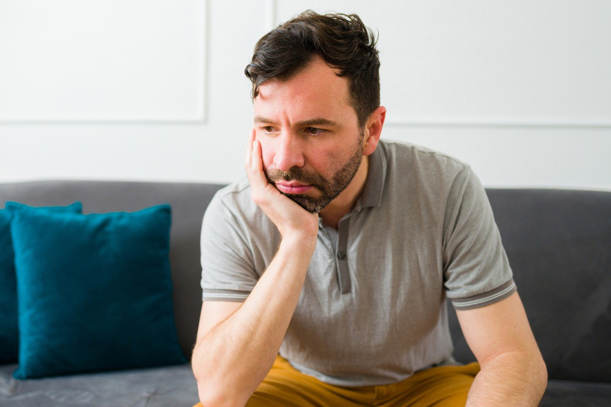 mężczyzna w wieku 35 plus z ciemnym zarostem siedzi na kanapie zmartwiony