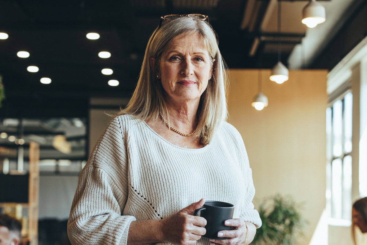 kobieta w wieku około 50 lat w jasnym swetrze stoi w biurze i lekko się uśmiecha