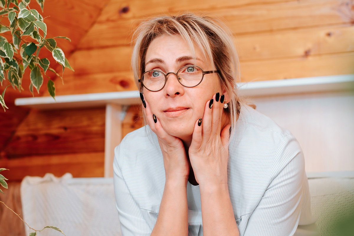 Adult lonely sad woman sitting on sofa and looking away, indoors. Middle aged caucasian woman wearing glasses and casual clothes alone at home. Loneliness, sadness, stress concept
