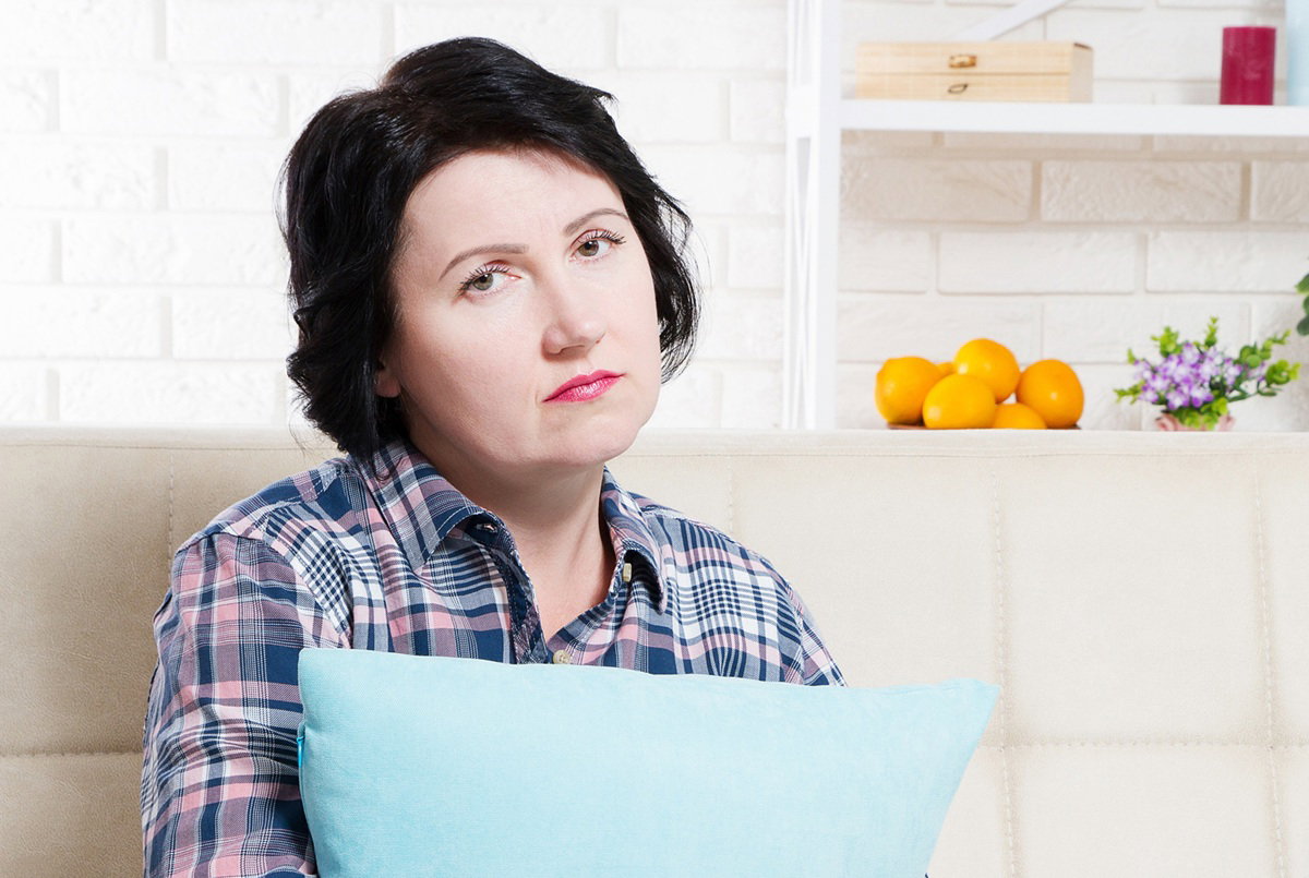 kobieta w wieku około 40 lat w koszuli w kratę siedzi na kanapie z poduszką smutna