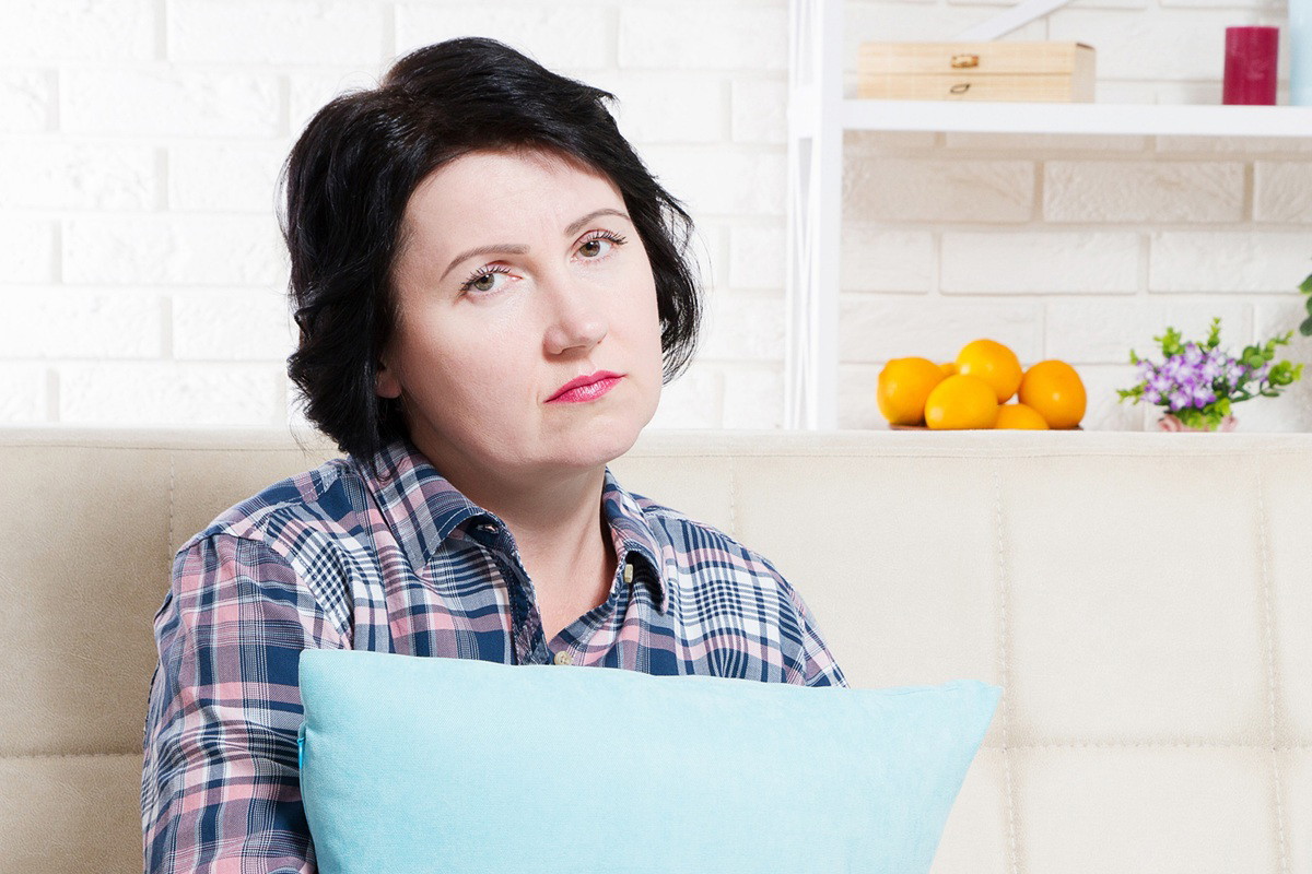 kobieta w wieku około 40 lat w koszuli w kratę siedzi na kanapie z poduszką smutna