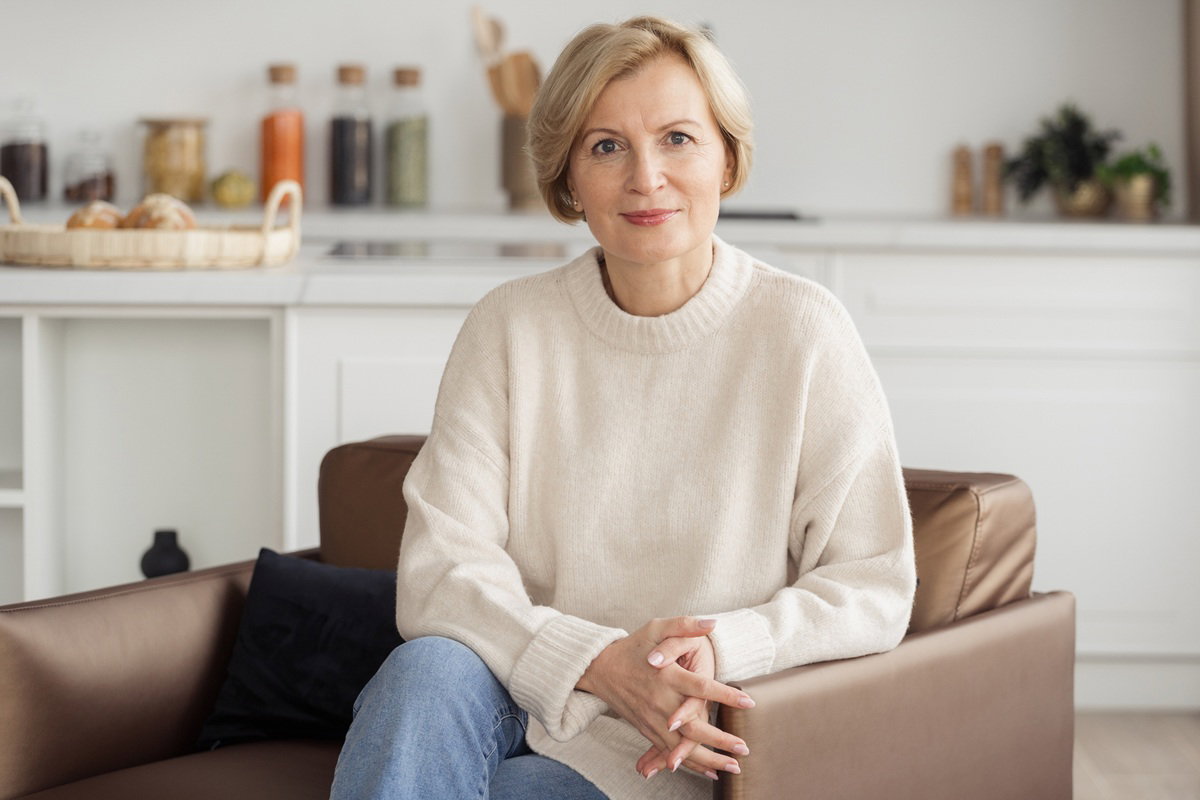kobieta w wieku 60 plus w jasnym swetrze siedzi na fotelu zamyślona i pogodna