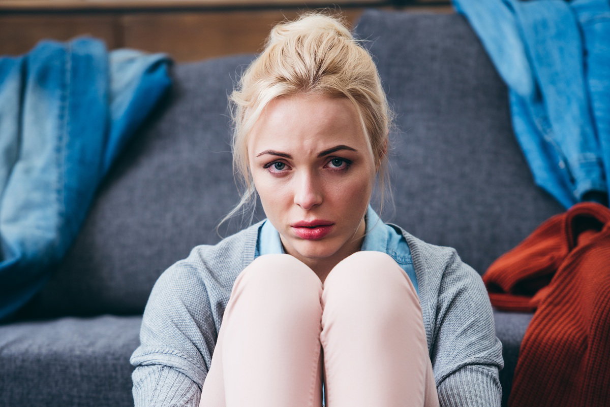 kobieta blondynka w wieku 30 plus siedzi na ziemi zmartwiona na tle kanapy z porozrzucanymi ubraniami