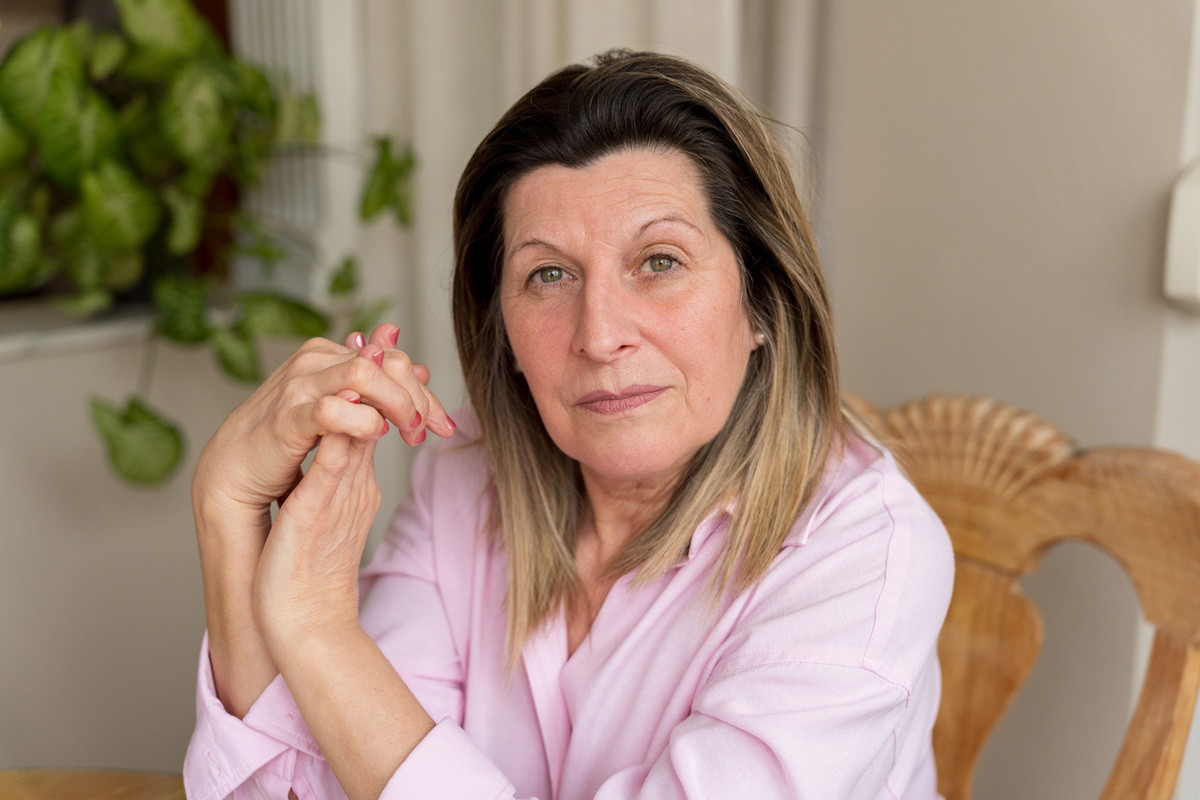 kobieta w wieku 65 plus w różowej koszuli siedzi na krześle z hardą miną
