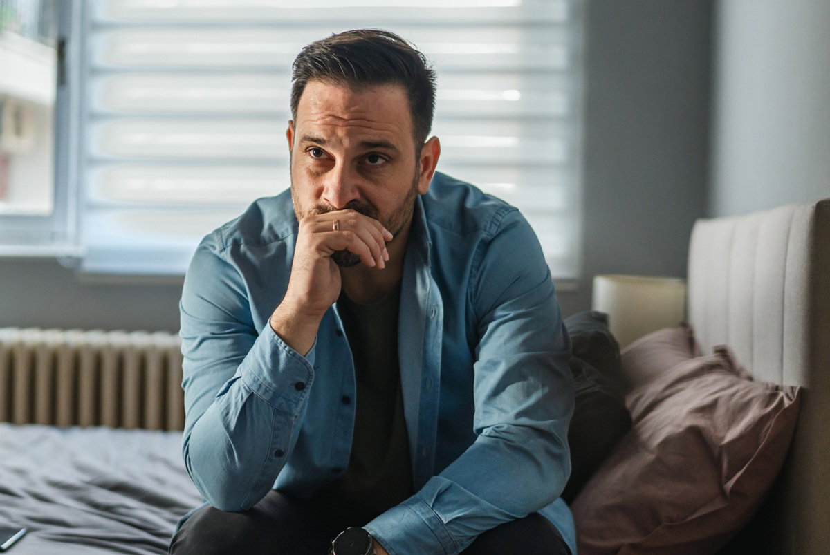 mężczyzna w wieku 35 plus w jasnej koszuli i z okularami w ręku siedzi na łóżku zmartwiony i zamyślony