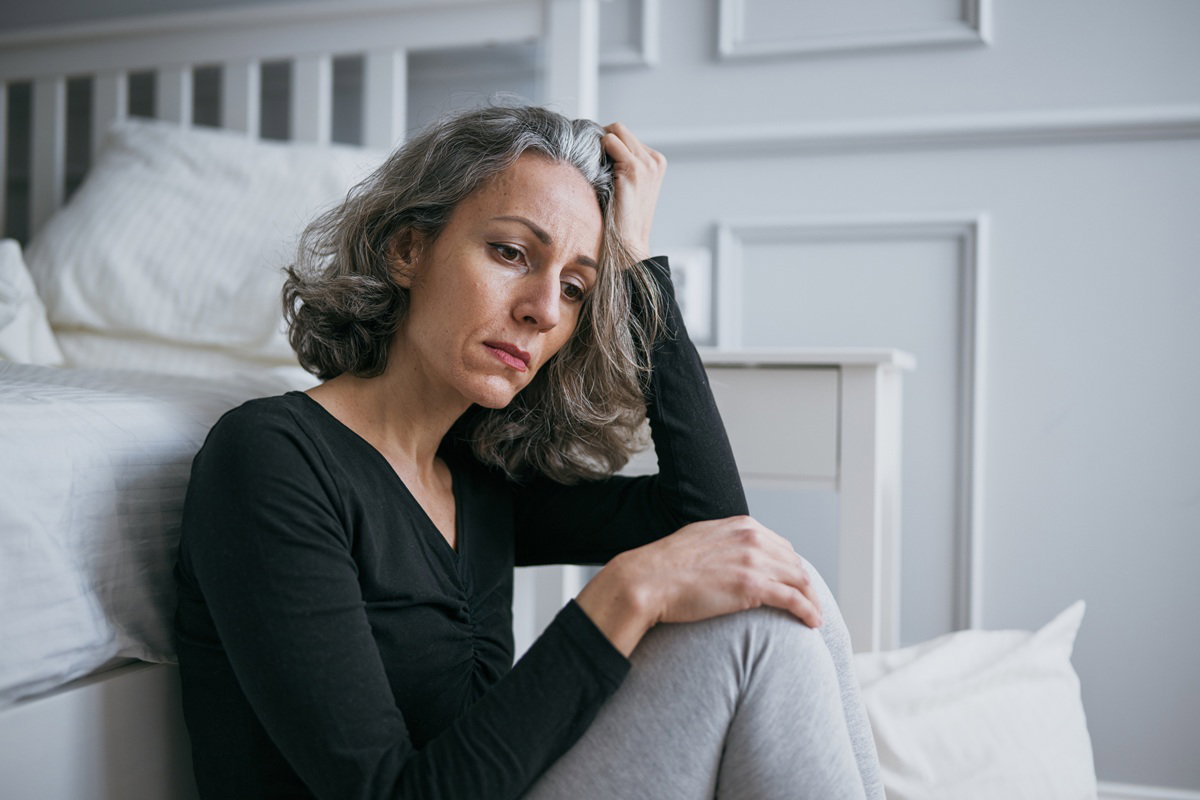 kobieta w wieku 40 plus siwa w czarnej bluzce siedzi na podłodze przy łóżku zamyślona i smutna
