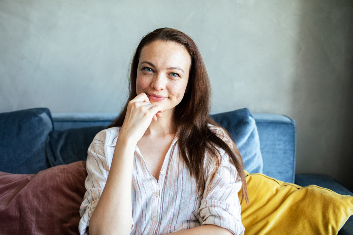 kobieta w wieku około 30 lat siedzi na łóżku z poduchami i się lekko uśmiecha