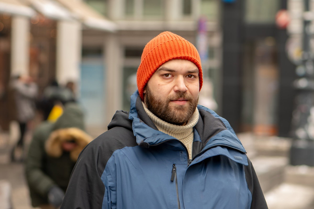 mężczyzna w wieku 35 plus z wąsami i brodą stoi na tle miasta w pomarańczowej czapce poważny