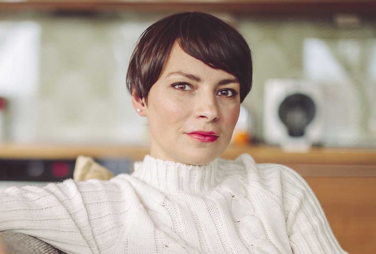 kobieta w wieku 40 plus z krótkimi ciemnymi włosami siedzi na kanapie na tle kuchni z poważną miną